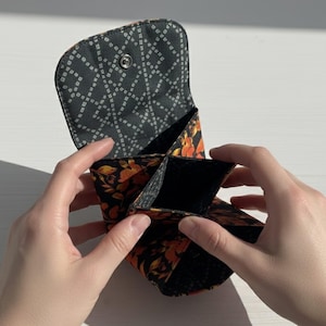 Puede incluir: Un peque&ntilde;o monedero abierto con un estampado floral negro y naranja. El interior est&aacute; dividido en m&uacute;ltiples compartimentos. La solapa del monedero es negra con un dise&ntilde;o geom&eacute;trico blanco y un cierre a presi&oacute;n.