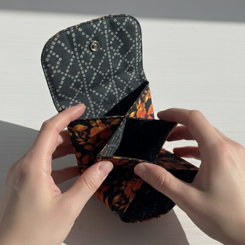 Puede incluir: Un peque&ntilde;o monedero abierto con un estampado floral negro y naranja. El interior est&aacute; dividido en m&uacute;ltiples compartimentos. La solapa del monedero es negra con un dise&ntilde;o geom&eacute;trico blanco y un cierre a presi&oacute;n.