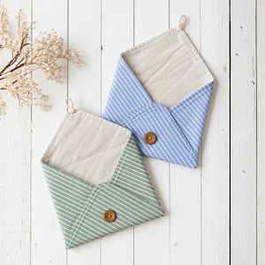 Op de afbeelding: Twee stoffen etuis in envelopstijl, één in lichtblauw met witte strepen en één in saliegroen met witte strepen. Elk etui heeft een beige voering, een enkele bruine houten knoopsluiting en een klein stoffen lusje aan de bovenhoek.
