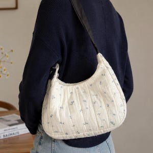 Könnte beinhalten: Eine weiße, gesteppte Hobo-Tasche mit kleinem blauem Blumenmuster und einem schwarzen Schulterriemen. Die Tasche hat eine Halbmondform mit vertikalen Steppnähten. Der Text "STEP BY STEP GUIDE BEGINNER FRIENDLY" steht unten.