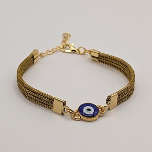 Peut inclure: Un bracelet composé de trois brins parallèles de cordon vert olive, fixés par des embouts en métal doré. Une breloque ronde bleu et blanc en forme d'œil est centrée, reliée à un fermoir mousqueton doré et une chaînette d'extension.