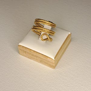 Könnte beinhalten: Ein goldfarbener Ring mit Spiralmuster, präsentiert auf einem kleinen weißen und Holzblock. Der Ring hat eine strukturierte Oberfläche und eine einzigartige Form. Der Ring ist ein Schmuckstück.