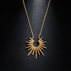 May include: A gold-toned necklace featuring a pendant shaped like a sunburst or a stylized horseshoe with radiating rays. The pendant is suspended from a delicate gold-toned chain, creating a bright, metallic focal point against the dark background.