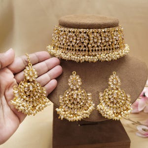 May include: A gold-coloured jewellery set featuring a choker necklace and matching earrings. The choker and earrings are adorned with clear, sparkling stones and small, white pearls. The earrings have a floral design and dangle with a cluster of pearls.