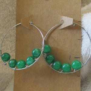 May include: A pair of silver hoop earrings with bright green beads. The beads are strung along the bottom of the hoops and secured with silver wire. The earrings are displayed on a brown earring card.