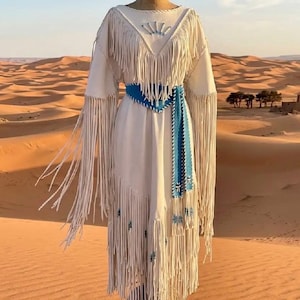 Puede incluir: Un vestido color crema con mangas largas y flecos. El vestido presenta un cinturón azul y detalles decorativos. Se exhibe en un maniquí en un paisaje desértico.