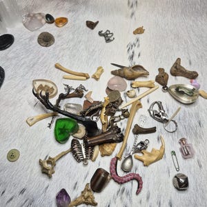 Könnte beinhalten: Eine Ansammlung verschiedener kleiner Objekte, darunter Knochen, Kristalle, eine Muschel und Metallanhänger, die auf einer weiß-grau gemusterten Oberfläche verstreut sind. Die Artikel variieren in Farbe und Textur und bilden ein einzigartiges Sortiment.