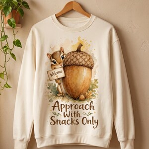 Könnte beinhalten: Ein cremefarbenes Sweatshirt mit Rundhalsausschnitt und einem Aquarell-Motiv eines Streifenhörnchens, das ein kleines Schild mit der Aufschrift Welcome und eine große Eichel hält. Darunter steht der Text Approach With Snacks Only in Braun.