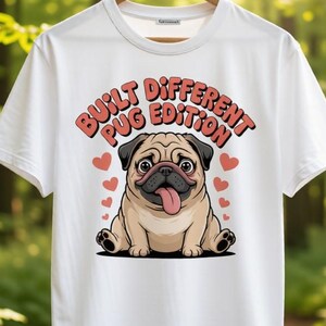 Könnte beinhalten: Weißes T-Shirt mit einer Cartoon-Mops-Illustration. Der Mops hat einen beigen Körper, ein schwarzes Gesicht und eine rosa Zunge. Der Text "BUILT DIFFERENT PUG EDITION" befindet sich über dem Mops, mit roten Herzen um das Design.