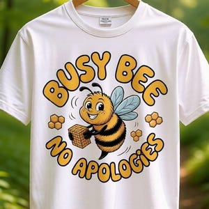 May include: White t-shirt with a cartoon bee graphic. The bee is yellow and black, holding a basket, with the words "Busy Bee No Apologies" in a playful font. The shirt is hanging on a wooden hanger.