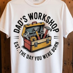 May include: White t-shirt featuring a toolbox graphic with tools and the text "DAD'S WORKSHOP EST. THE DAY YOU WERE BORN" in a circular design. The toolbox is open, displaying various tools.