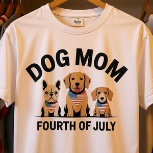 May include: White t-shirt featuring the text "DOG MOM" in black. Below are three cartoon dogs wearing red, white, and blue bandanas. The words "FOURTH OF JULY" are printed beneath the dogs.