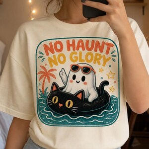 Può includere: Una t-shirt color crema con una stampa grafica di un fantasma bianco con occhiali da sole arancioni e un salvagente nero a forma di gatto che galleggia nell'acqua blu. Il testo "NO HAUNT NO GLORY" è stampato in arancione sopra il fantasma.
