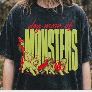 May include: A charcoal grey acid-wash t-shirt featuring a graphic print with the text "dog mom of MONSTERS" in red and lime green. The illustration depicts a person in a witch hat walking three dogs in costumes, with "spooky szn" printed below.