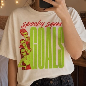 May include: A cream-colored short-sleeve t-shirt featuring a graphic print with the red text "spooky squad" above the large green text "GOALS". The graphic includes three stylized figures wearing sunglasses and necklaces in red and green tones.