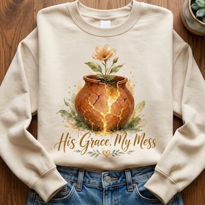 May include: A beige crewneck sweatshirt featuring a graphic of a cracked terracotta pot with a blooming flower inside. The text "His Grace, My Mess" is printed below the pot in a brown script font, accompanied by small green leaves and a heart.