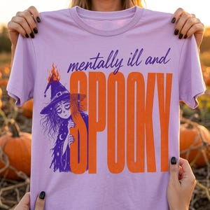 May include: A light purple short-sleeve t-shirt featuring a graphic of a witch wearing a hat with flames. The text reads "mentally ill and" in purple script above the large, bold, orange, vertical block letters spelling "SPOOKY".