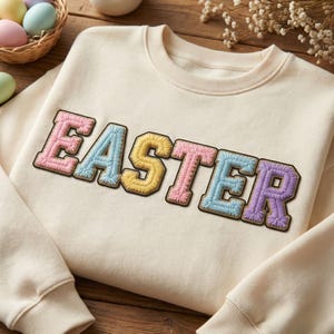 May include: Cream-colored sweatshirt with the word "EASTER" embroidered in pastel colors. The letters are pink, blue, yellow, pink, and purple, with a brown border. The sweatshirt has a crew neck and long sleeves, perfect for Easter.