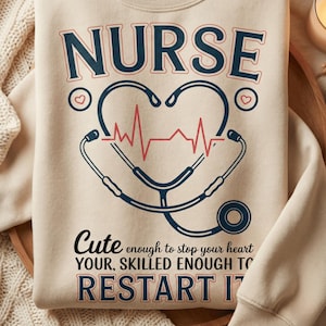 May include: Beige sweatshirt with a navy blue and red graphic. The graphic features the word "NURSE" above a heart-shaped stethoscope with a red heartbeat line. Below the stethoscope, the text reads "Cute enough to stop your heart, your skilled enough to restart it."