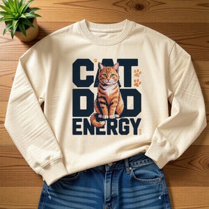 Peut inclure: Sweat-shirt crème avec le texte "CAT DAD ENERGY" en lettres majuscules bleu marine. Une illustration détaillée d'un chat tigré est centrée au-dessus du mot "ENERGY". Deux empreintes de pattes orange sont en haut à droite.