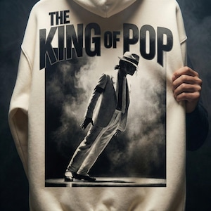 May include: Cream-colored hoodie featuring a black and white image of a person in a suit and fedora, with the text "THE KING OF POP" above. The image depicts a famous lean pose. The hoodie is made of a soft material.