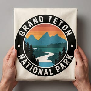 Pode incluir: Camiseta creme com um gráfico circular do Parque Nacional Grand Teton. O design inclui montanhas, um rio, árvores e o texto "GRAND TETON NATIONAL PARK" em uma borda preta com estrelas brancas.