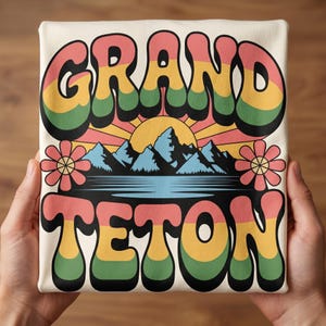 May include: Cream-colored t-shirt featuring the words "GRAND TETON" in a retro, colorful font. The design includes a mountain range, sun, and flowers, evoking a vintage travel aesthetic. The shirt is held by hands.