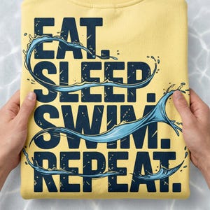 Może przedstawiać: Ż&oacute;łta bluza z granatowym napisem "EAT. SLEEP. SWIM. REPEAT.". Niebieskie grafiki z rozpryskami wody dodają dynamiki i atrakcyjności wizualnej.
