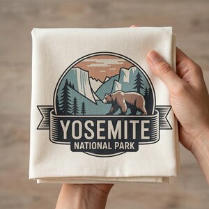 May include: Cream-colored kitchen towel featuring a Yosemite National Park design. The illustration includes a bear, mountains, and the text "YOSEMITE NATIONAL PARK" in a banner. The towel is folded and held by a person.