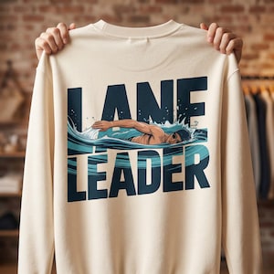 Może przedstawiać: Kremowa bluza z granatową grafiką pływaka w wodzie, z napisem "LANE LEADER". Pływak ma na sobie czepek i okulary pływackie. Bluza jest trzymana przez dwie ręce.