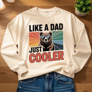 以下が含まれることがあります： 「LIKE A DAD JUST COOLER」の文字とサングラスをかけたクマのグラフィックがプリントされたクリーム色のスウェットシャツ。クマはカラフルなストライプの背景に配置されています。スウェットシャツはブルージーンズと組み合わせています。