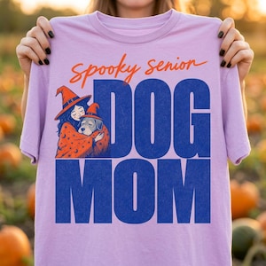 May include: A light purple t-shirt featuring the orange text "spooky senior" above the large blue text "DOG MOM". The graphic includes an illustration of a person wearing a witch hat and a star-patterned cloak, hugging a dog also wearing a witch hat.