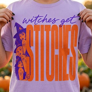 May include: A light purple t-shirt featuring a graphic print with the text "witches get STITCHES" in purple and orange. The design includes three illustrated figures wearing witch hats, each with a small skull tattoo on their arm.