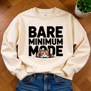 May include: Cream-colored sweatshirt with the text "BARE MINIMUM MODE" in bold black letters. A cartoon cat with brown and tan stripes is nestled beneath the text. The sweatshirt is paired with blue jeans.