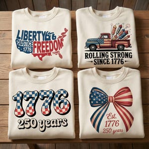 May include: Four cream-colored sweatshirts with patriotic designs. One features a map of the USA with "Liberty, Justice, Freedom, 1776, Years." Another shows a truck with fireworks and "Rolling Strong Since 1776." The others have "1776 250 years" and a bow with "Est. 1776 250 years."
