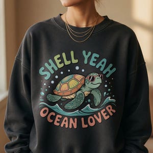 May include: Dark gray sweatshirt with a colorful graphic of a cartoon turtle wearing sunglasses and the words "SHELL YEAH OCEAN LOVER". The turtle is swimming in blue water with bubbles. The sweatshirt is a casual style.