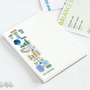 May include: A white notepad with a colorful graphic of charts and graphs, with the name "JUDY WILLIAMS" printed at the bottom. A white pen with red accents lies next to the notepad. Financial and business-related stationery.