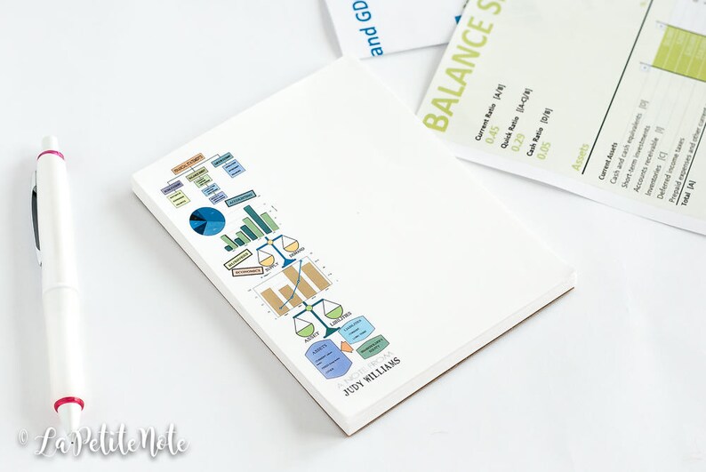 May include: A white notepad with a colorful graphic of charts and graphs, with the name "JUDY WILLIAMS" printed at the bottom. A white pen with red accents lies next to the notepad. Financial and business-related stationery.