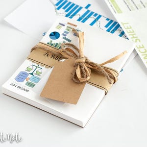 May include: A white notepad bundle tied with twine and a blank tag. The notepad has a printed label with the text "La Petite Note" and "JUDY WILLIAMS". A pen and financial documents are also in the image.