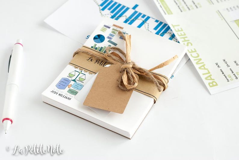 May include: A white notepad bundle tied with twine and a blank tag. The notepad has a printed label with the text "La Petite Note" and "JUDY WILLIAMS". A pen and financial documents are also in the image.