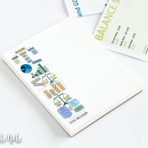 May include: White notepad with colorful business charts and graphs. A white pen with red accents is on the left. The notepad has the name "JUDY WILLIAMS" printed on it. A document with the words "BALANCE SHEET" is in the background.