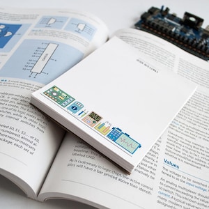 May include: A close-up of a white notepad with a colorful illustration of various electronic components, including a circuit board, a multimeter, an oscilloscope, and a soldering iron. The notepad has the text "La Petite Note" printed on the bottom left corner.