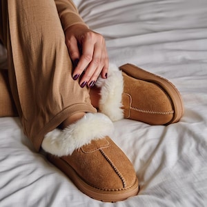 Puede incluir: Zapatillas de ante marrón con forro de piel sintética color crema. Las zapatillas tienen una suela gruesa marrón y están diseñadas para uso en interiores. La imagen muestra las zapatillas en una cama.