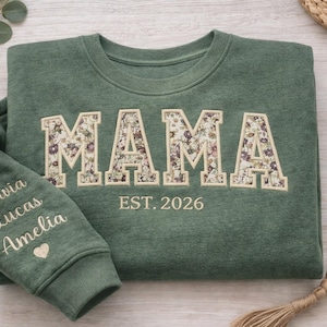 May include: A green sweatshirt with the word "MAMA" in floral print letters. The sleeve has embroidered names and a heart. The text "EST. 2026" is below the word "MAMA".