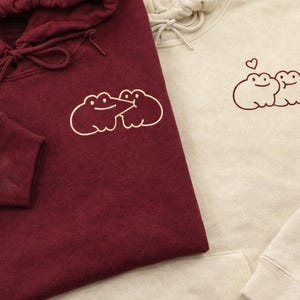 May include: Two hoodies, one burgundy and one cream, are displayed. Each features a frog design. The burgundy hoodie has a frog graphic on the front and a small design on the sleeve. The cream hoodie has a frog design with a heart above it.