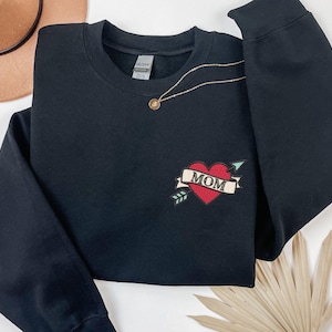 May include: A black long-sleeve crewneck sweatshirt featuring an embroidered design on the left chest. The embroidery depicts a red heart pierced by a light blue arrow, with a beige banner draped across the heart containing the black text MOM.