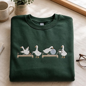 May include: A dark forest green crewneck sweatshirt featuring an embroidered graphic of four white geese performing various Pilates exercises on equipment, including using a reformer, a stability ball, and a ring, with orange beaks and feet.