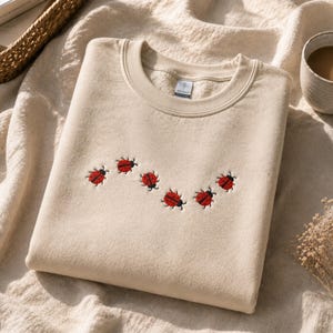 May include: A beige crewneck sweatshirt featuring an embroidered design of six small red ladybugs with black spots arranged in a gentle, wavy line across the center of the chest. The garment has a ribbed collar and a standard long-sleeve construction.