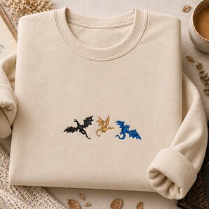 May include: Cream-coloured sweatshirt with embroidered dragon silhouettes in black, gold, and blue. The crew neck sweatshirt has long sleeves and a relaxed fit. The design features three stylized dragons.