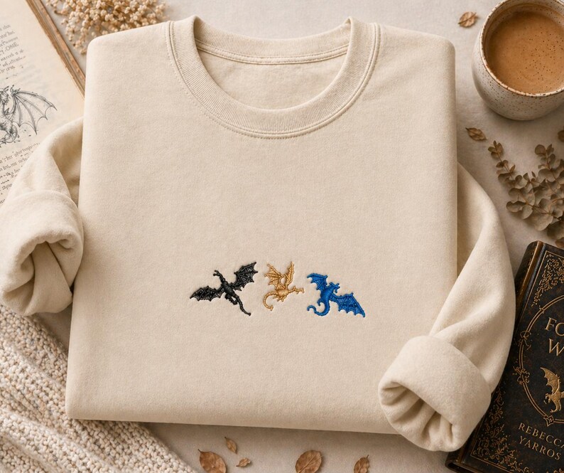 May include: Cream-coloured sweatshirt with embroidered dragon silhouettes in black, gold, and blue. The crew neck sweatshirt has long sleeves and a relaxed fit. The design features three stylized dragons.